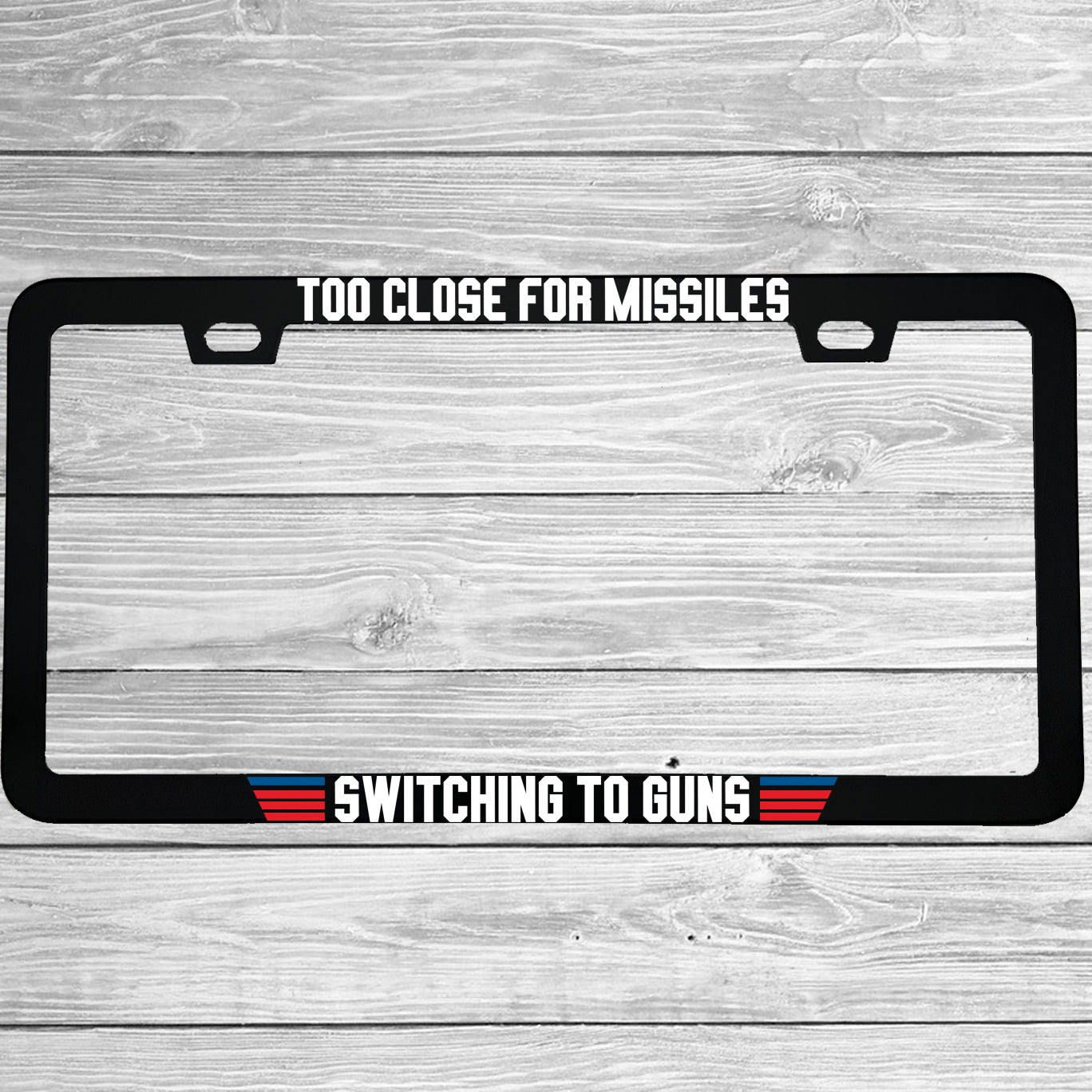 Close-up of "Too Close For Missiles Switching To Guns" license plate frame showing fade-proof print quality