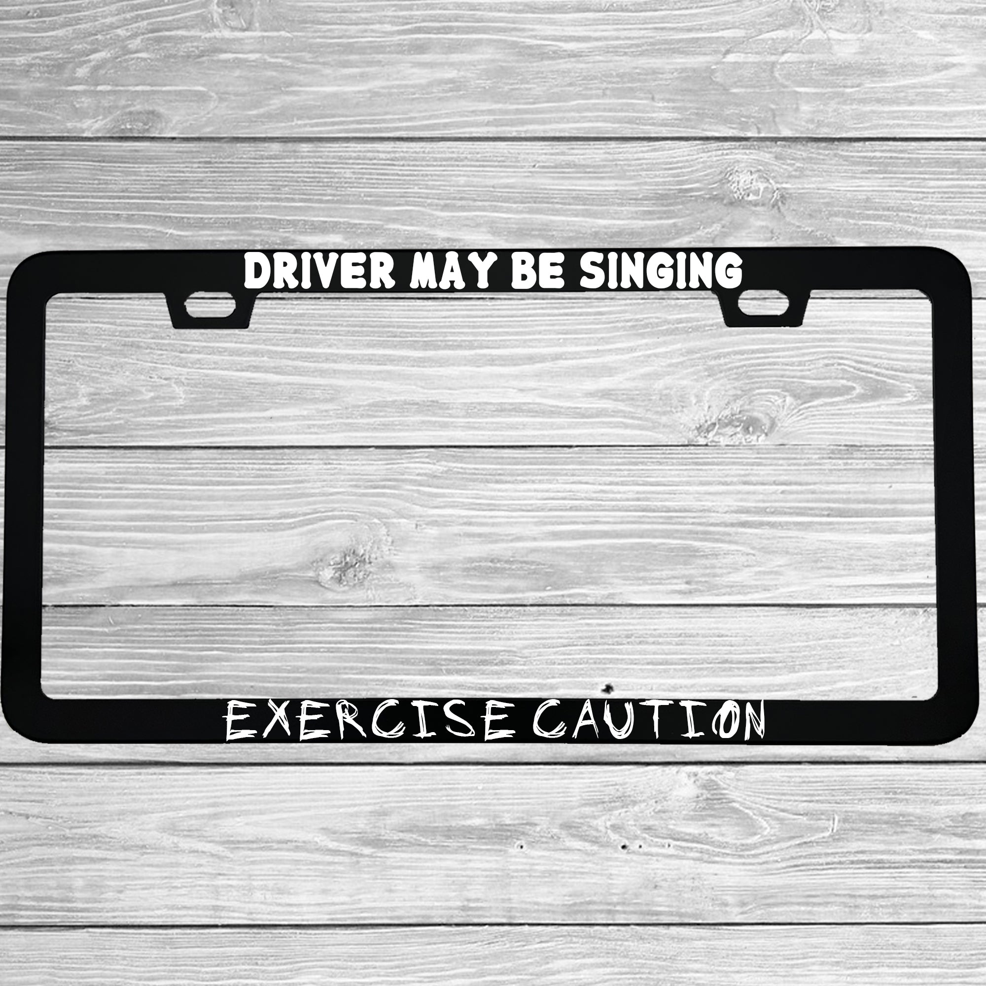 Close-up of "Driver May Be Singing Exercise Caution" license plate frame showing fade-proof print quality